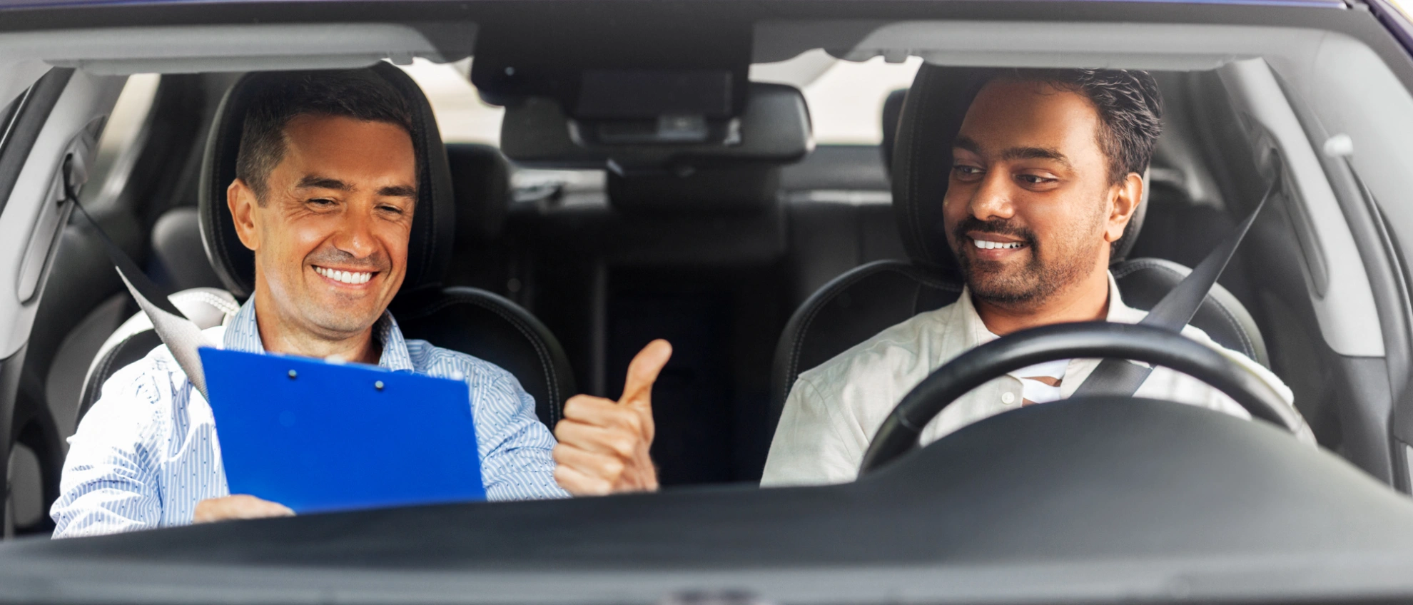 Theorie trifft Praxis Fahrlehrer erklärt dem Schüler im Auto etwas mit einem Klemmbrett in der Hand, beide lachen