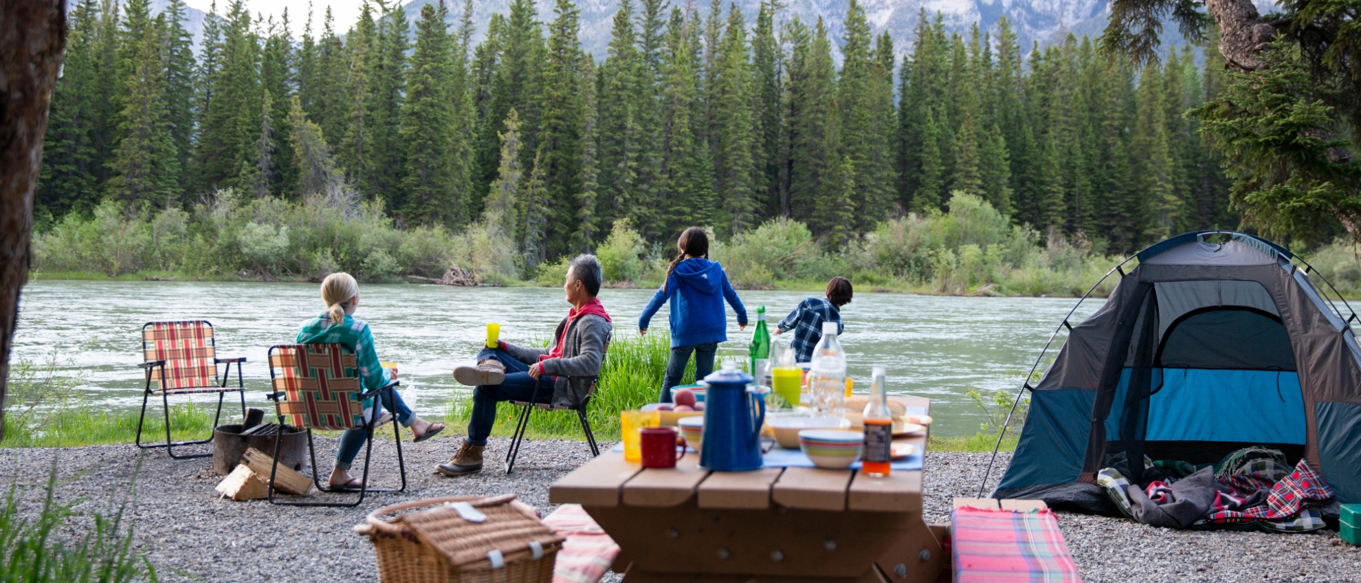 Camping mit der Familie Familie mit Kindern beim Zelten auf einer Wiese vor einem Wald, Tische und Stühle aufgebaut