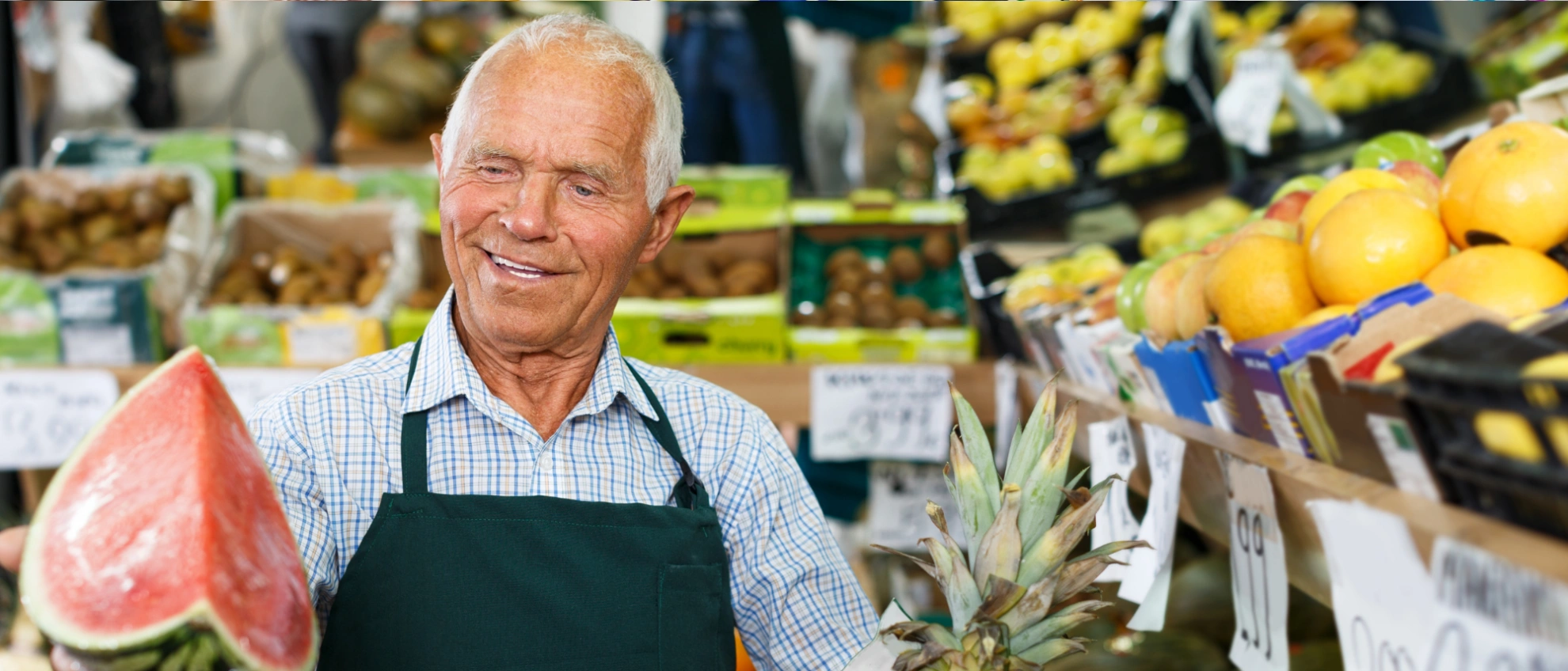 Älterer Mann mit Schürze steht an einem Obst- und Gemüsestand und hält ein Netz mit Kartoffeln