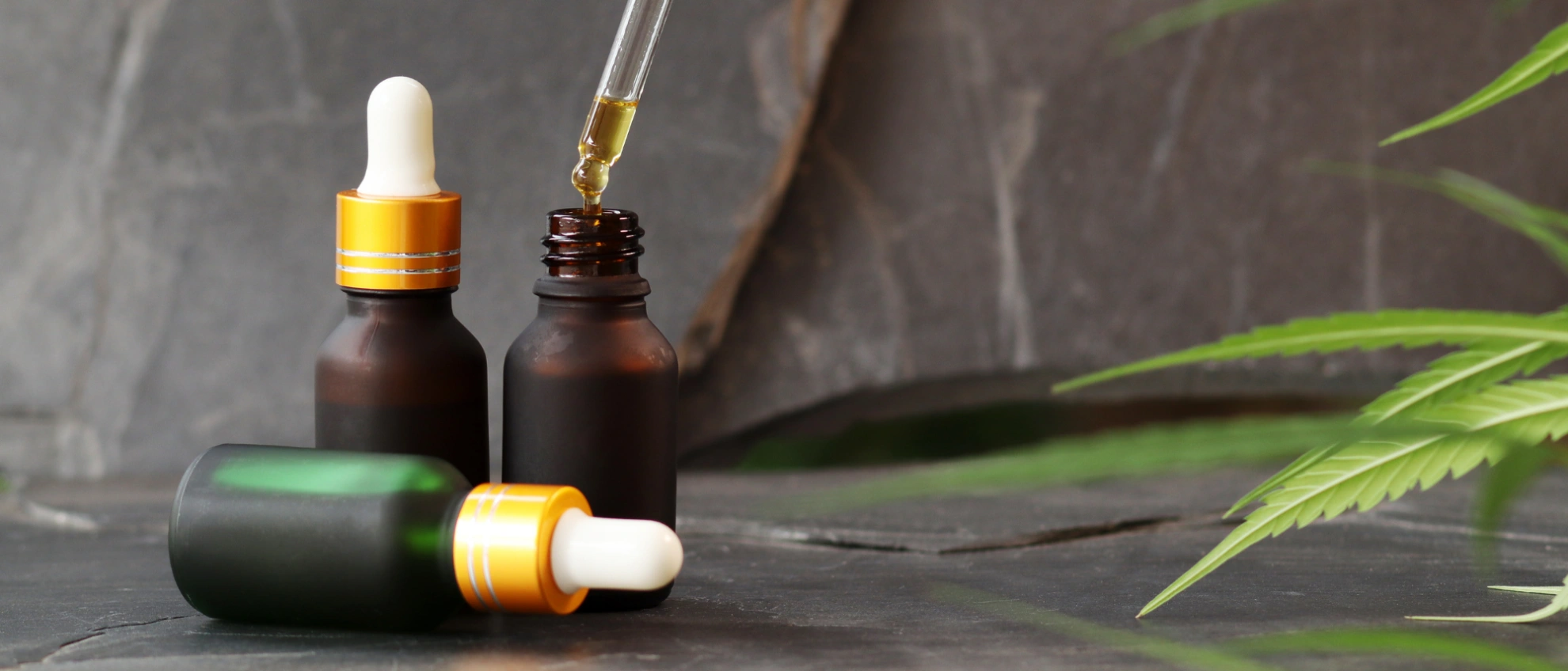 Braunes CBD-Fläschchen mit Pipette, Tropfen werden neben Hanfblatt auf Marmoroberfläche entnommen