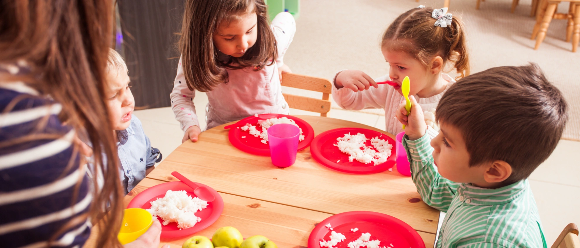 Kinder sitzen an einem Tisch und essen zusammen mit rotem KindergeschirrBällebad-Spaß in der Kita