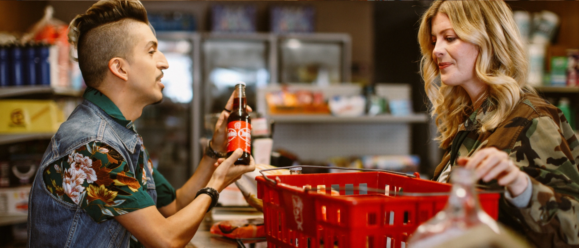Mann reicht einer Kundin ein Produkt über eine rote Einkaufskiste in einem gut gefüllten Laden