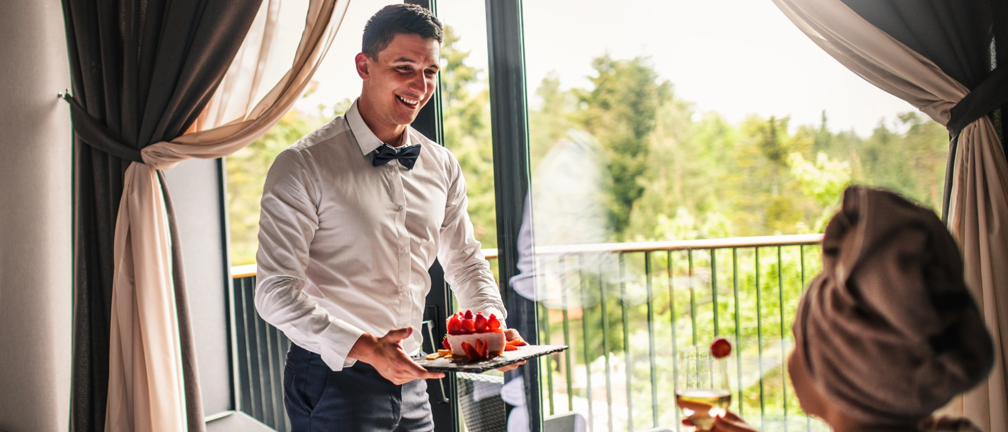 Hotelangestellter serviert einem Gast auf dem Balkon Frühstück mit Aussicht ins Grüne