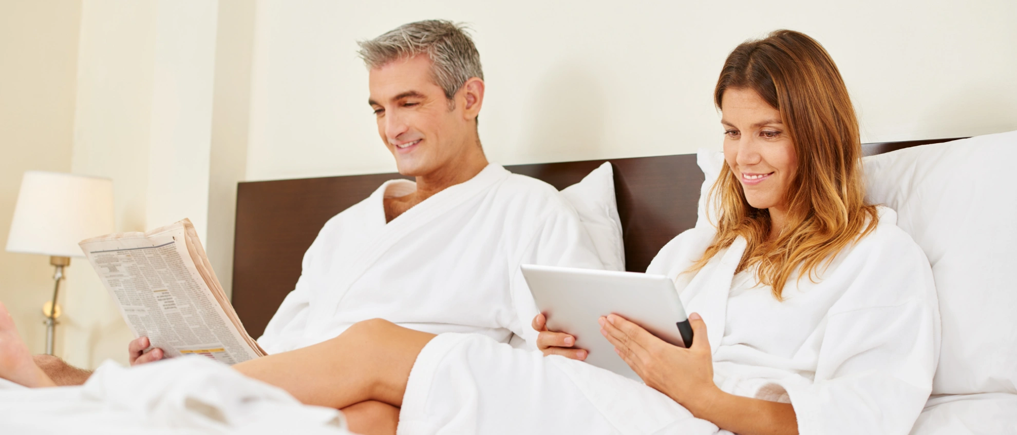 Mann und Frau im Hotelbett, beide im Bademantel, nutzen Handy und lesen Zeitung