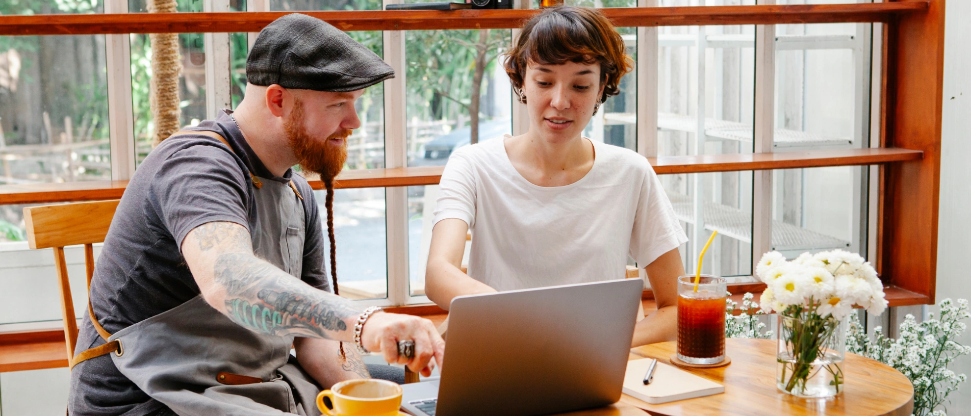 Zwei Personen sitzen mit einem Laptop an einem Fensterplatz in einem gemütlichen Café