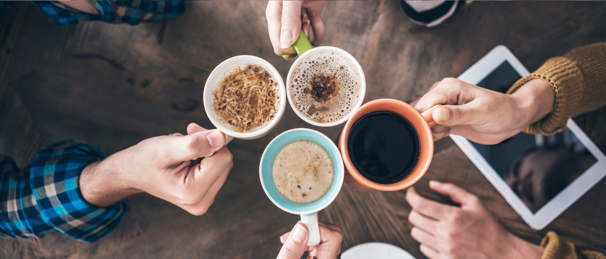 Mehrere Hände halten Tassen mit verschiedenen Kaffeegetränken über einem Holztisch