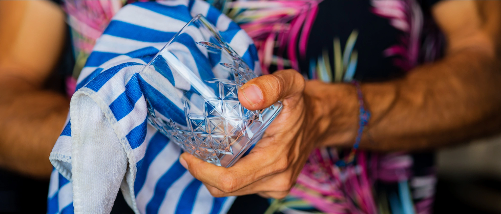 Barkeeper trocknet ein edles Glas mit einem blau-weiß gestreiften Tuch vor bunter Hemd-Kulisse