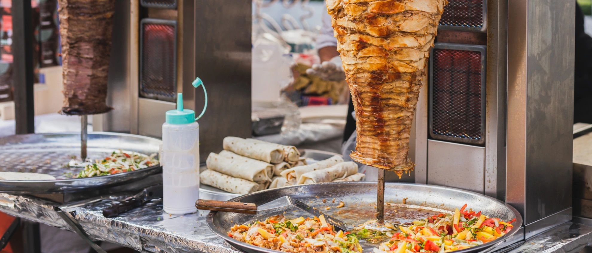Dönerfleisch wird an mehreren vertikalen Spießen gegrillt, daneben Schalen mit Salat und Soße