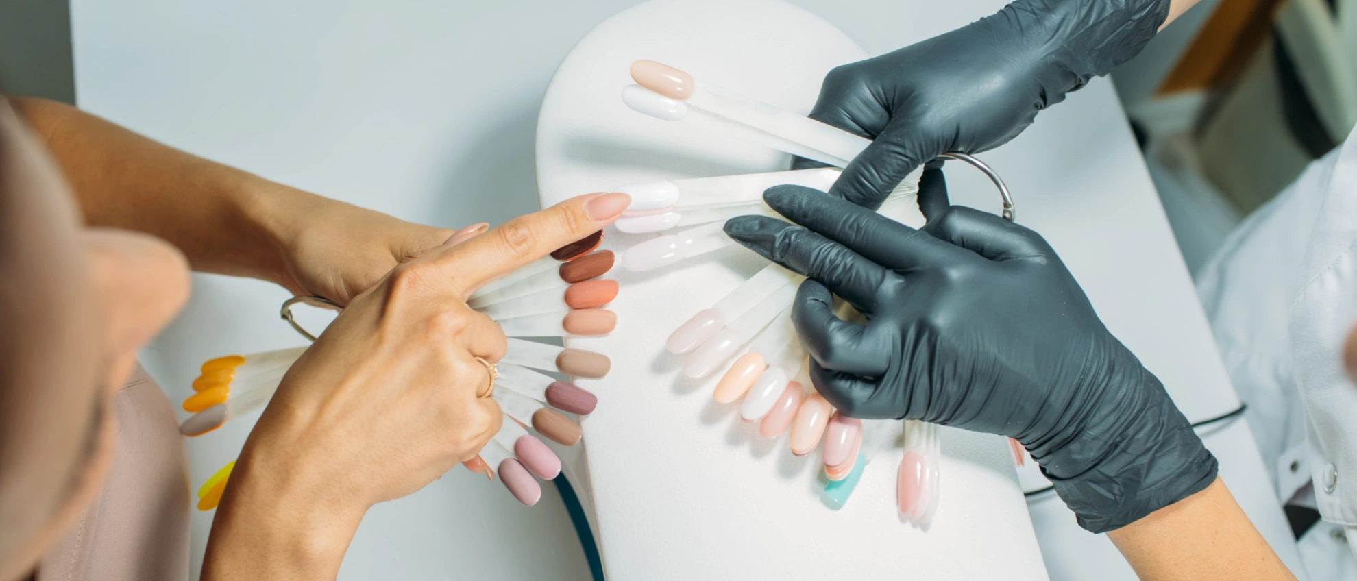 Zwei Hände halten eine Farbmuster-Palette mit künstlichen Fingernägeln, eine Kundin zeigt auf ihren Wunschton