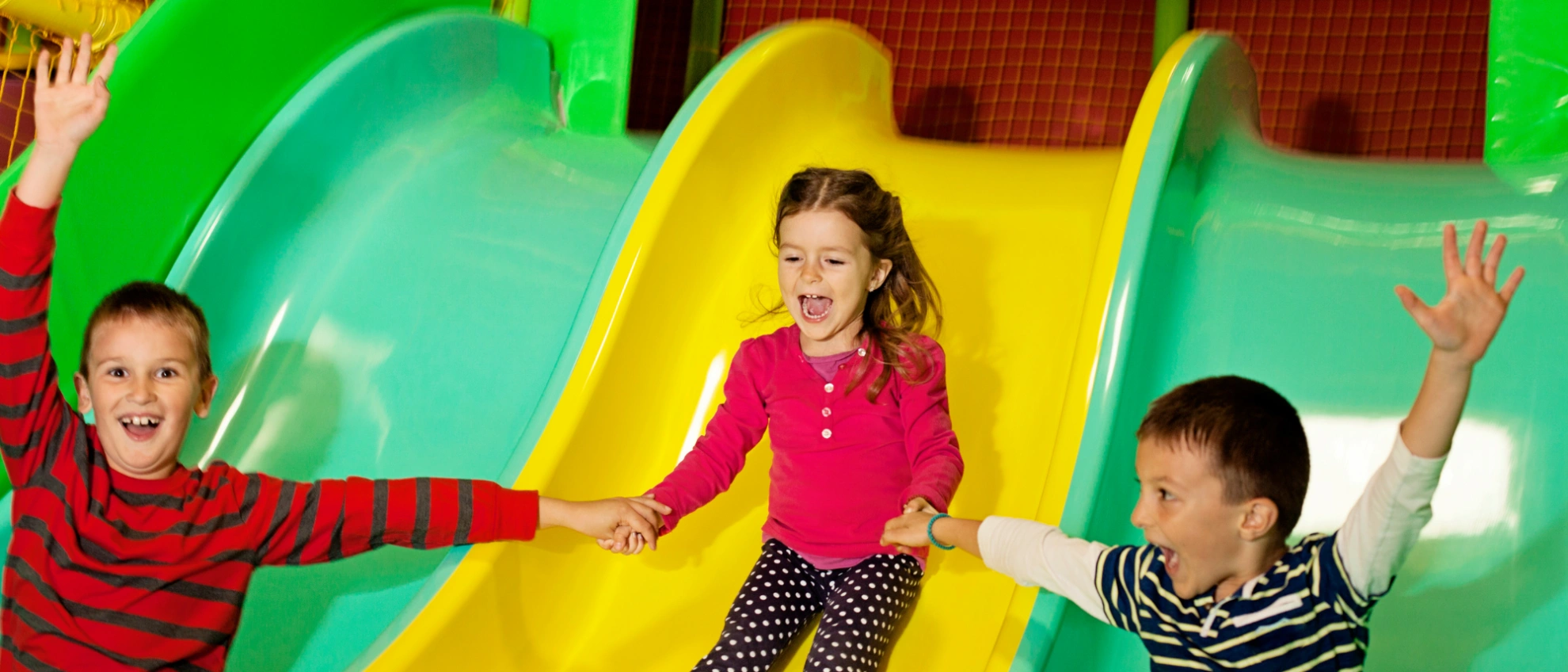 Drei Kinder rutschen lachend eine gelbe und grüne Wellenrutsche hinunter