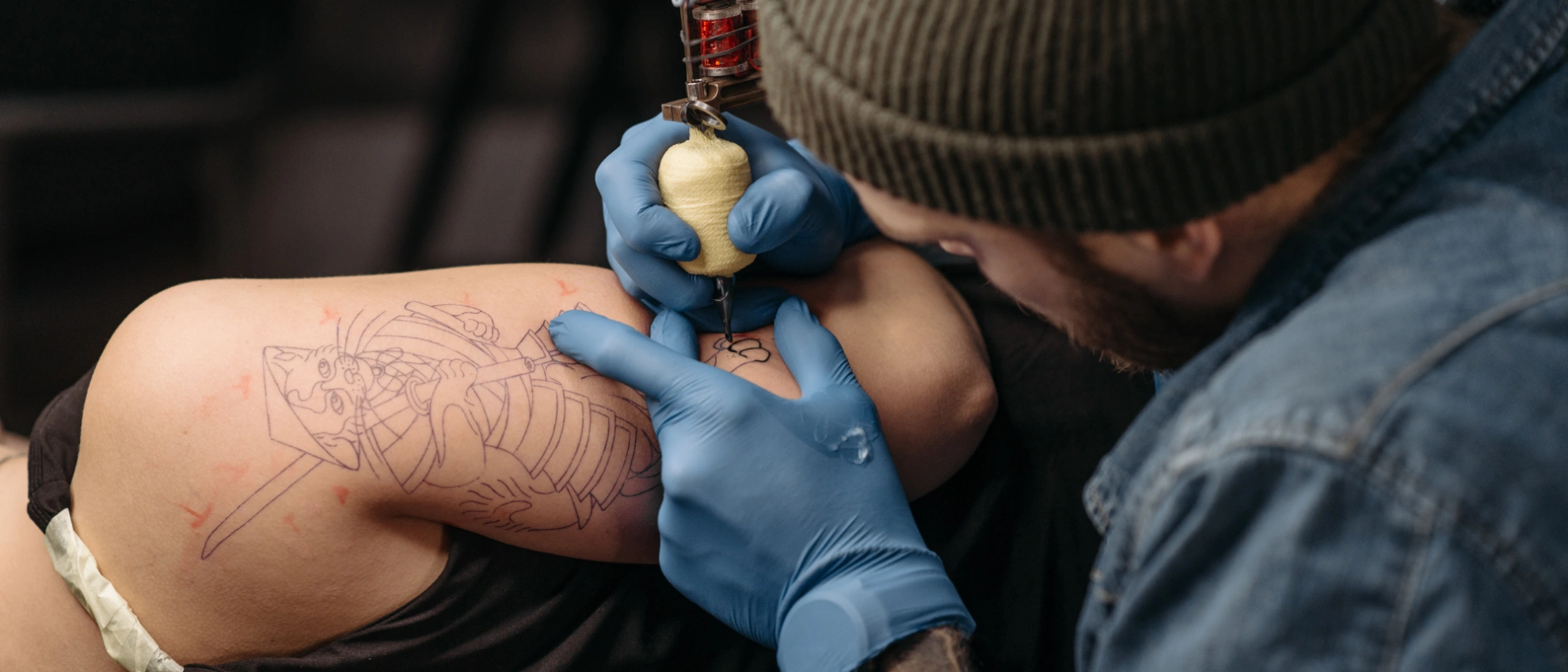 Frisches Tattoo in Arbeit – Linien und Schattierungen auf dem Oberarm Tätowierer mit blauen Handschuhen arbeitet an einem Oberarmtattoo mit violetter Vorzeichnung