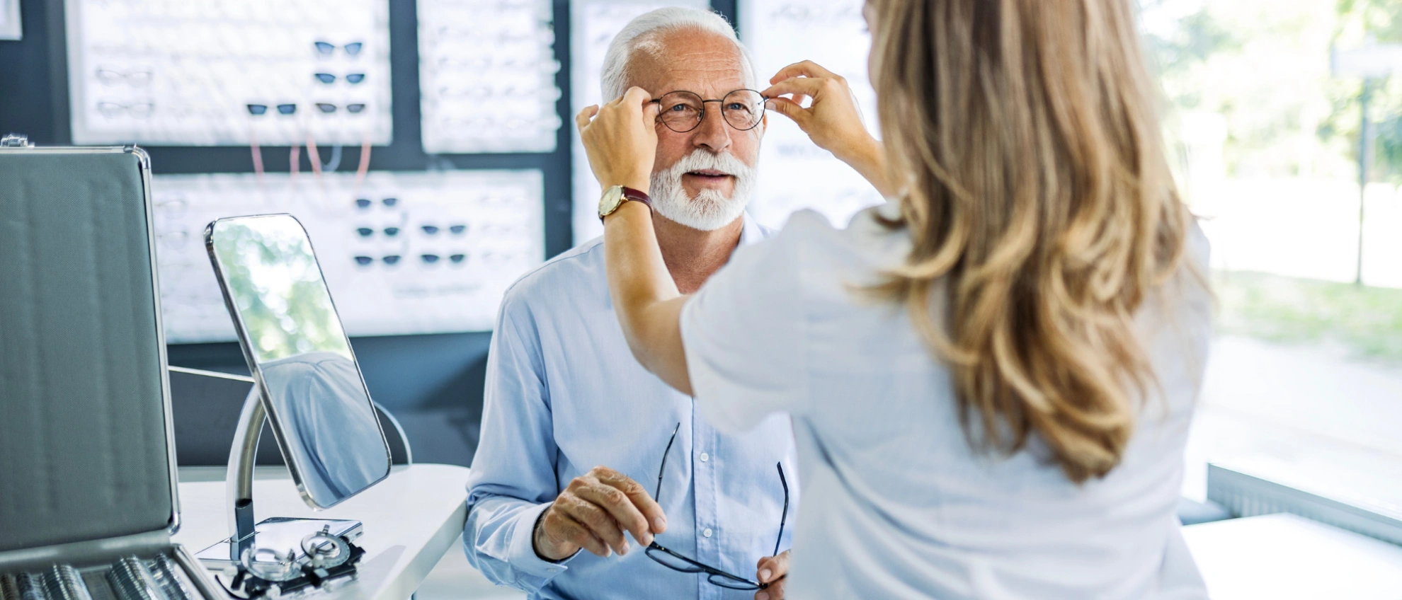 Optikerin berät älteren Kunden bei einem Sehtest, beide sitzen vor modernen Diagnosegeräten im hellen Raum