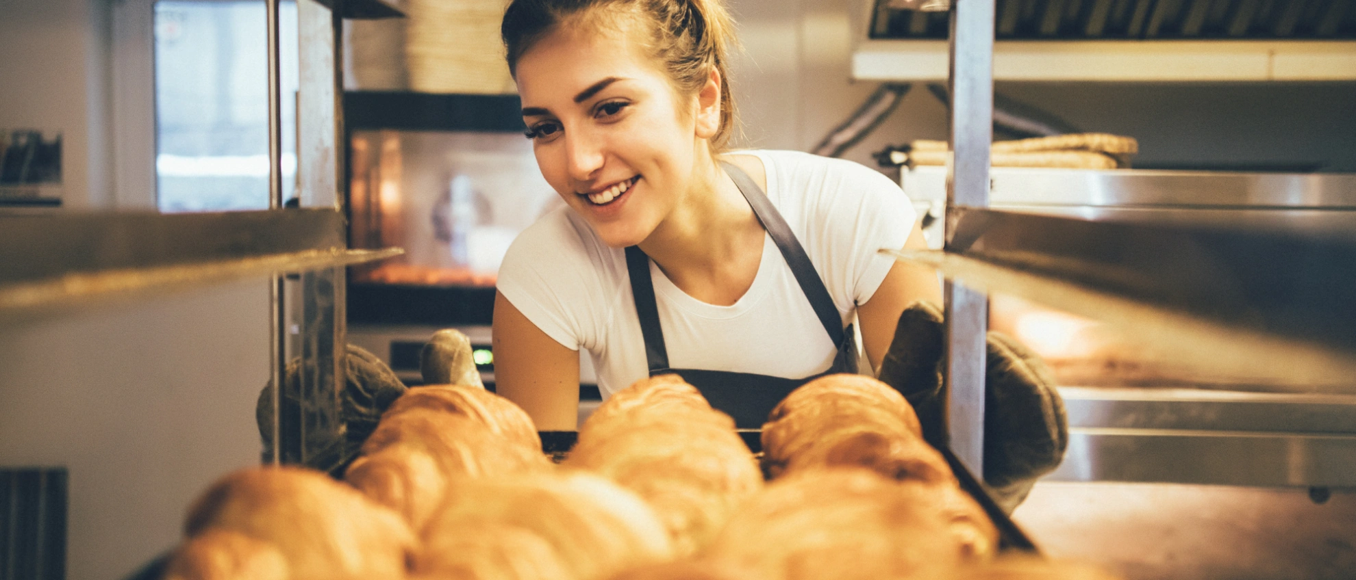 Ofenfrische Brötchen und Backfreude in der Bäckerei Bäckerin lächelt, während sie Brote aus dem Ofen entnimmt, im Vordergrund goldbraune Backwaren