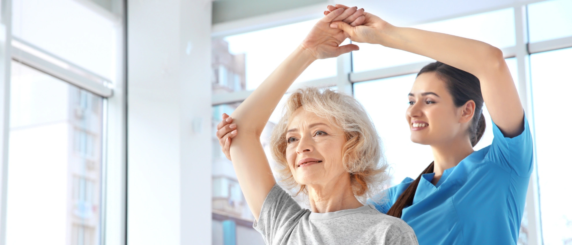Physiotherapeut hilft einer älteren Frau bei einer Arm-Streckübung im hellen Behandlungsraum
